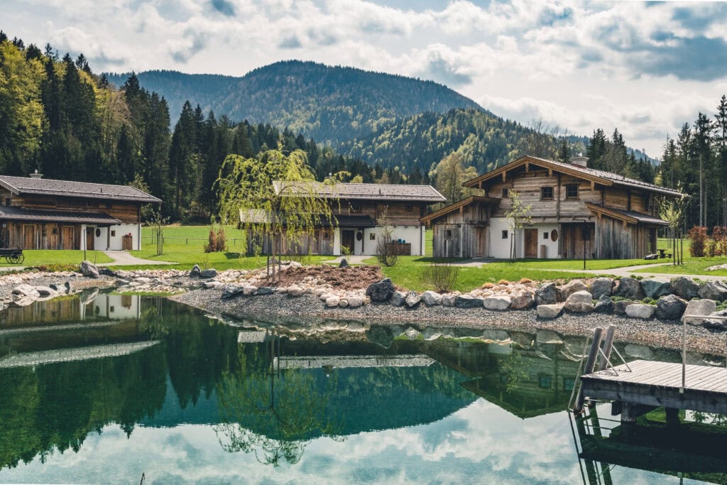 Naturschwimmteich mit Holzsteg und traditionellen Chalets vor Alpenpanorama, Luxusresort Bayern