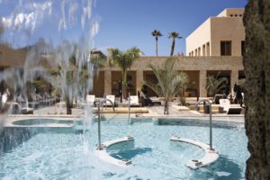 Pool mit Wasserfall, Liegen und Palmen, modernes Hotel im Hintergrund, sonniger Tag.