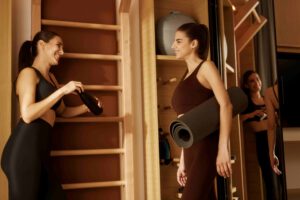 Zwei Frauen in Yoga- oder Fitnesskleidung im Fitnessraum, eine hält eine Wasserflasche, die andere Y.